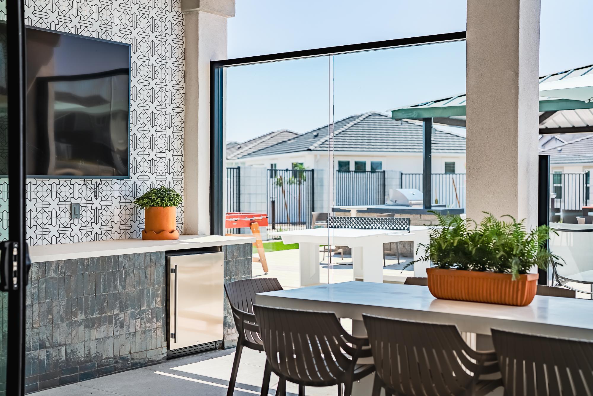 Modern outdoor patio with dining table, TV, potted plants, and open glass doors leading to a sunny backyard.