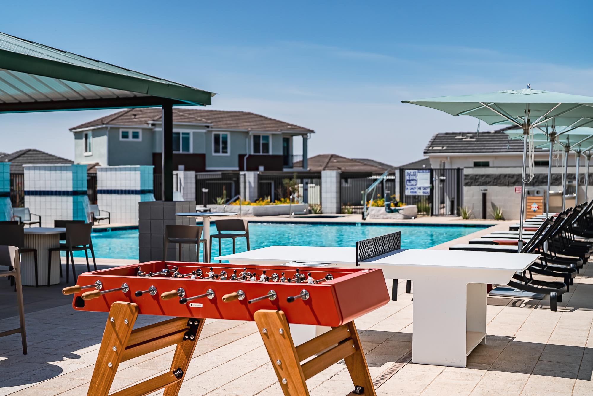 A red foosball table beside a pool with lounge chairs, tables, and houses in the background under a clear sky.